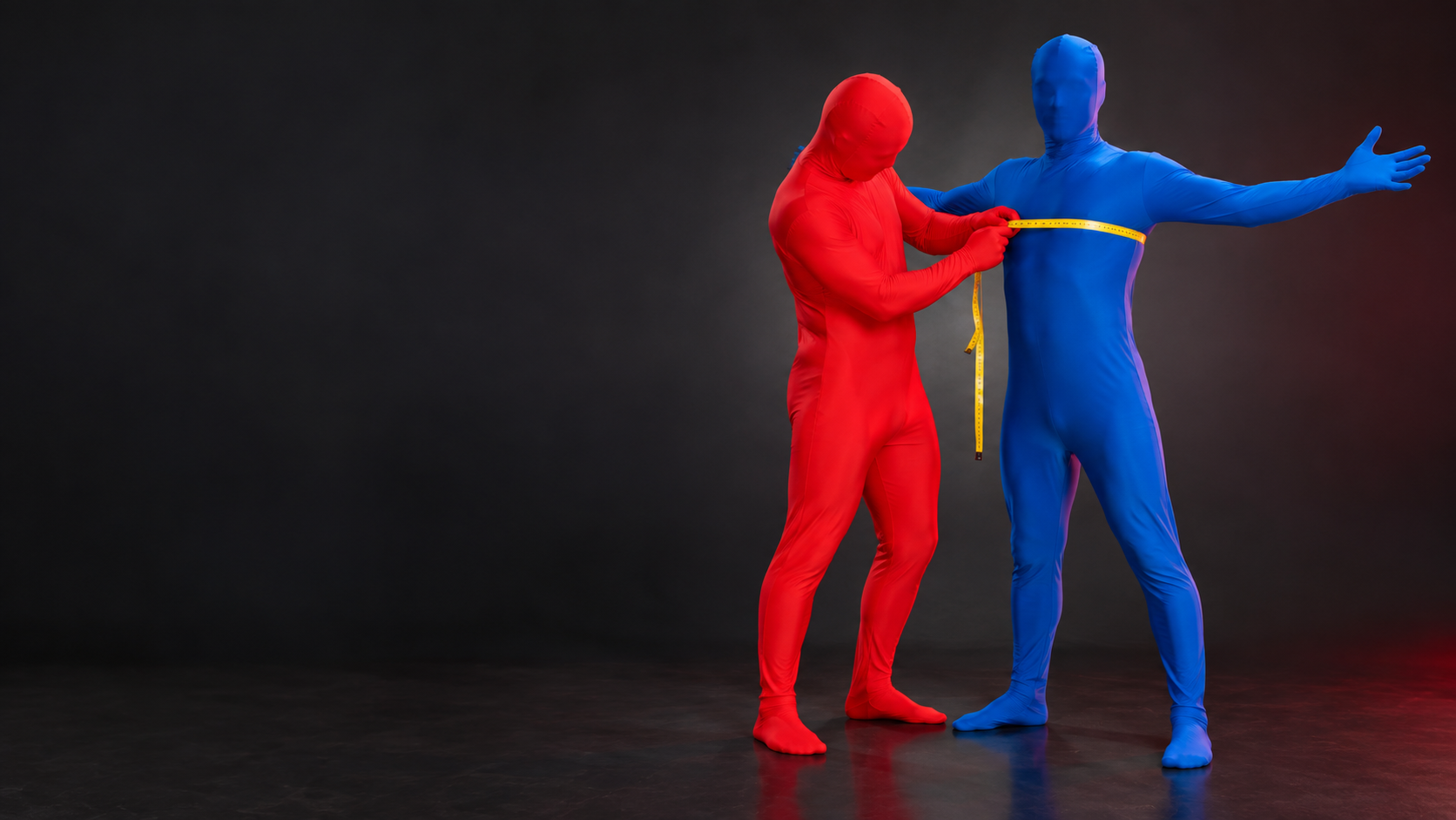 Two people in red and blue Morphsuits, one measuring the other's chest with a tape measure