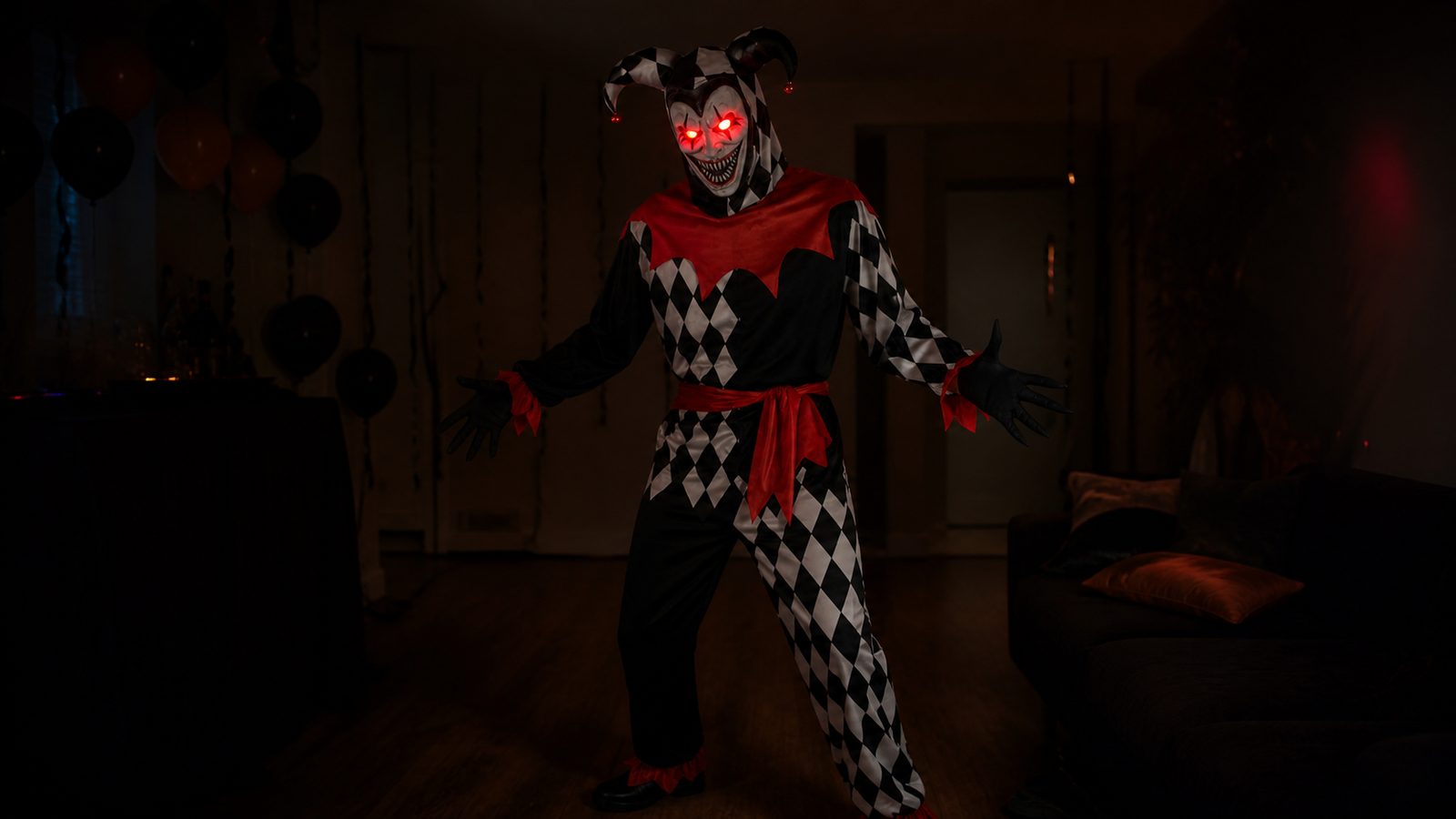 Person wearing the light-up jester mask in a dim room, the mask's red eyes glowing clearly against the diamond-print hood