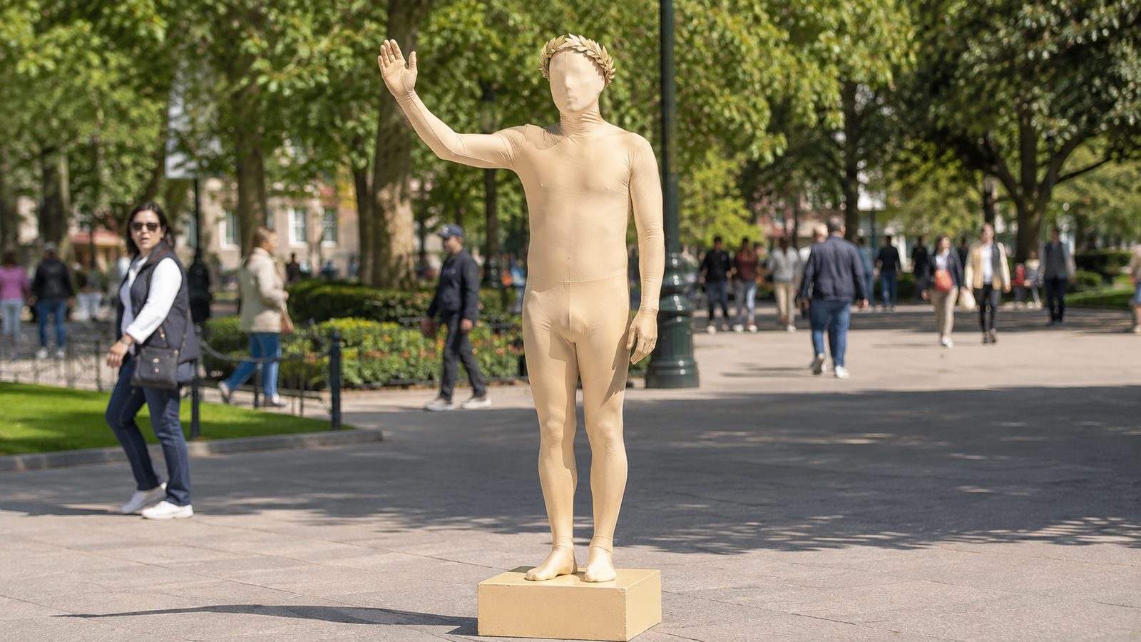Person in a pale champagne-gold full-body Morphsuit wearing a laurel wreath, standing rigid on a small plinth like a living statue