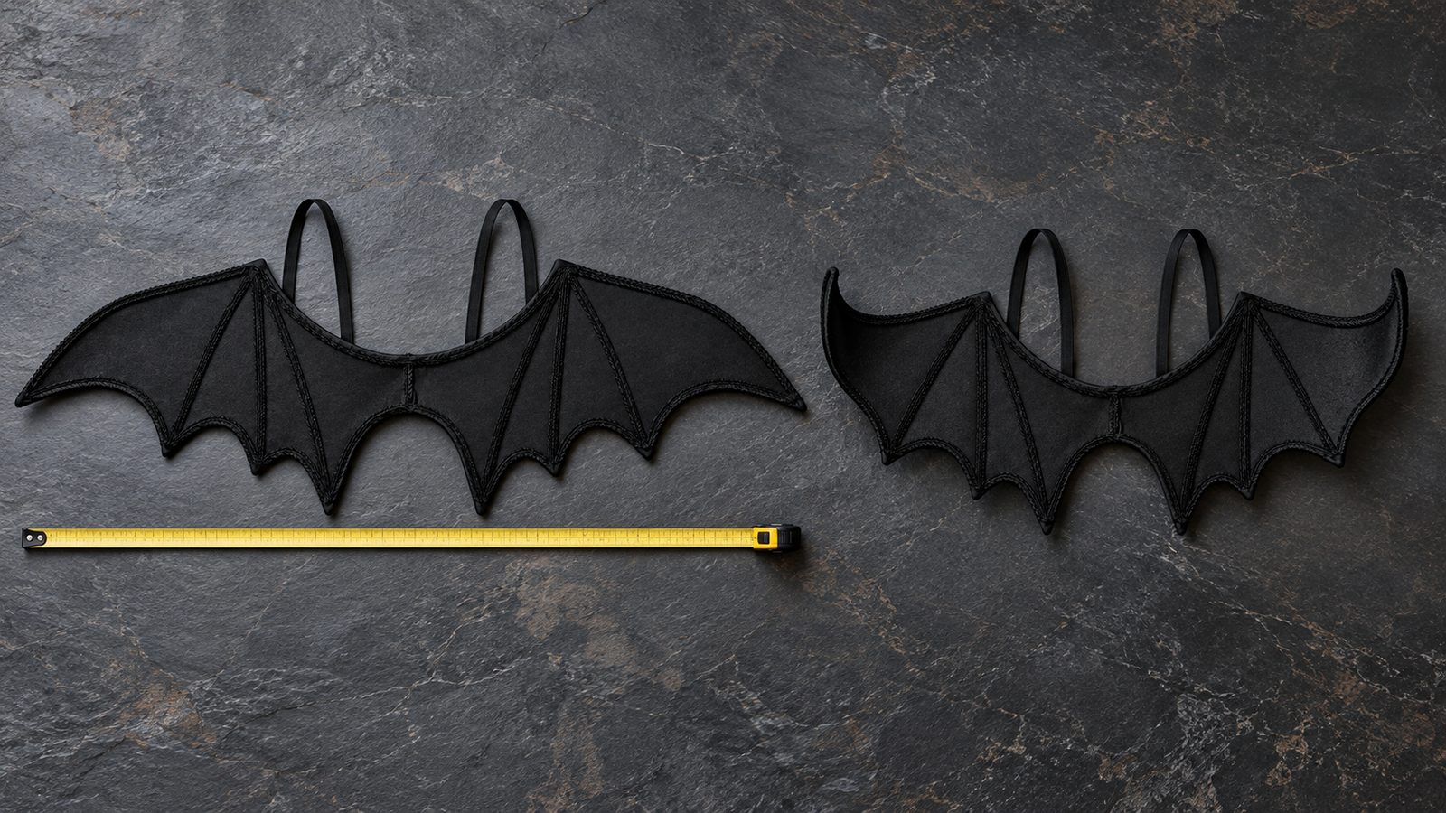 Two pairs of black felt bat wings with raised rib lines and elastic shoulder straps on a dark table, one opened flat with a tape measure along its width and one shaped into a deeper swoop