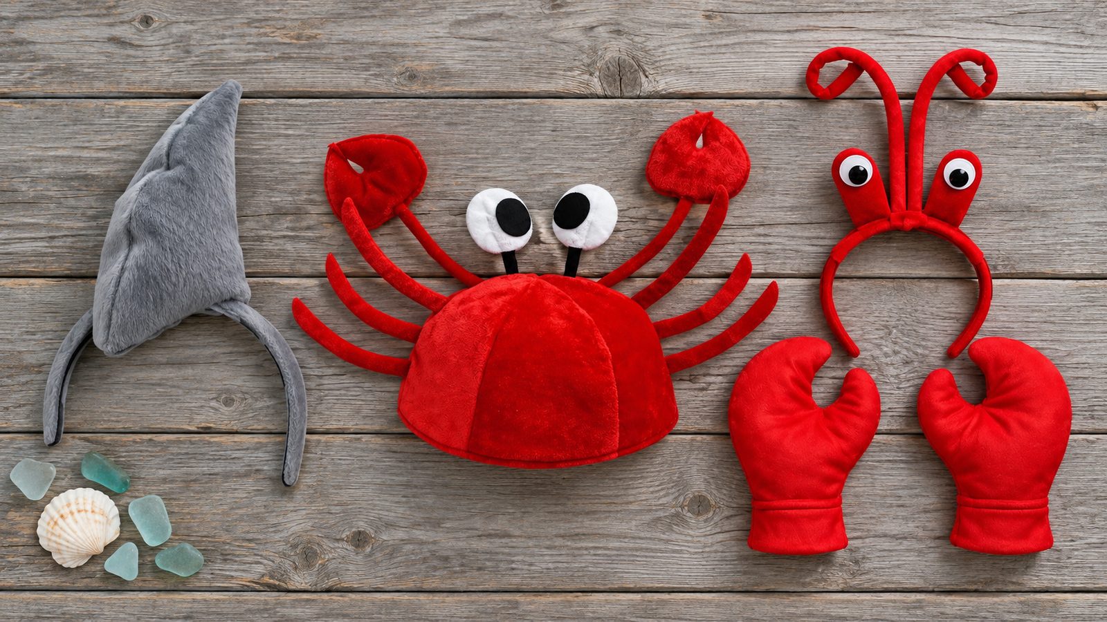 Flat-lay on weathered boards of a gray shark fin headband, a red crab hat with raised claws and eyes, and a red antenna headband beside a pair of red claw mitts